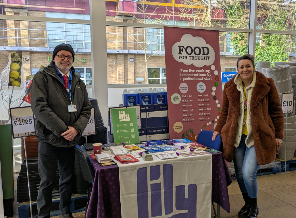 Tesco_Downham_010325 Lorcan and Becky from Careline and LILY standing in the foyer next to a table of leaflets and care alarms.
